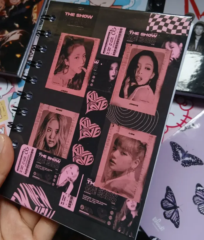 Agenda anillada de tapa dura negra, con un collage de fotografías y recortes en tonos rosados, sostenida sobre otras libretas ilustradas