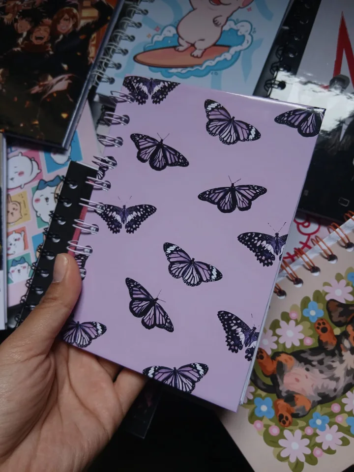 Agendita anillada lila con mariposas sobre otras libretas ilustradas