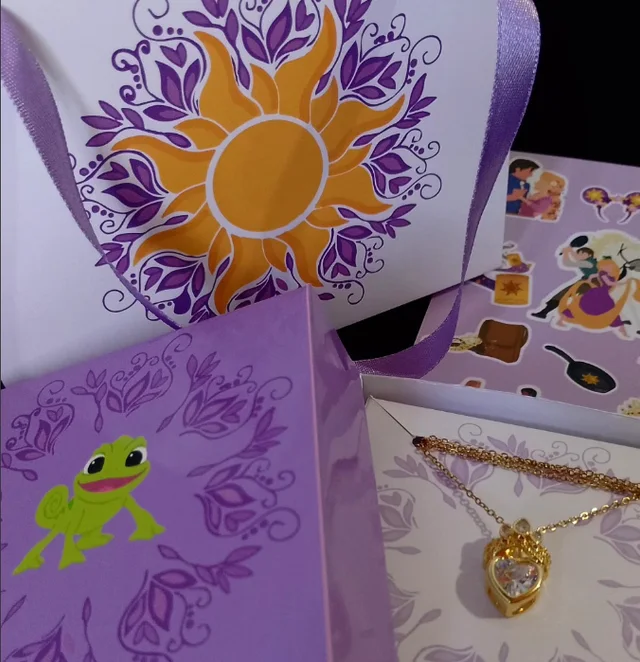Cadenita dorada con un dije en forma de corazón, presentada sobre el interior ilustrado de una caja, junto a otras cajas en tonos lilas con dibujos