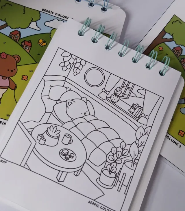 Cuaderno anillado abierto en una lámina para colorear con el dibujo de un gato durmiendo en una habitación, apoyado sobre otros cuadernos con tapas ilustradas a color